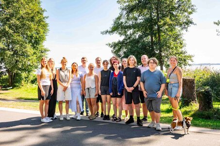 Eine Gruppe junger Menschen steht im Sommer vor einem See und schaut fröhlich in die Kamera.